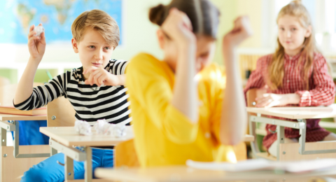 Children in classroom with one child throwing rolled up paper balls at another's back