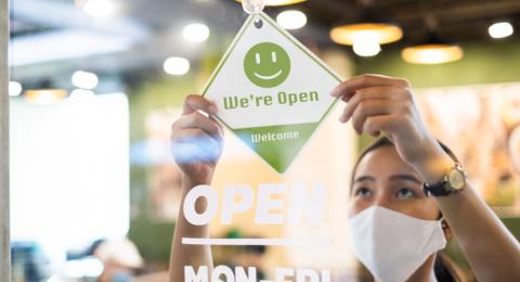 person in face mask hanging up an "open" sign in a store window